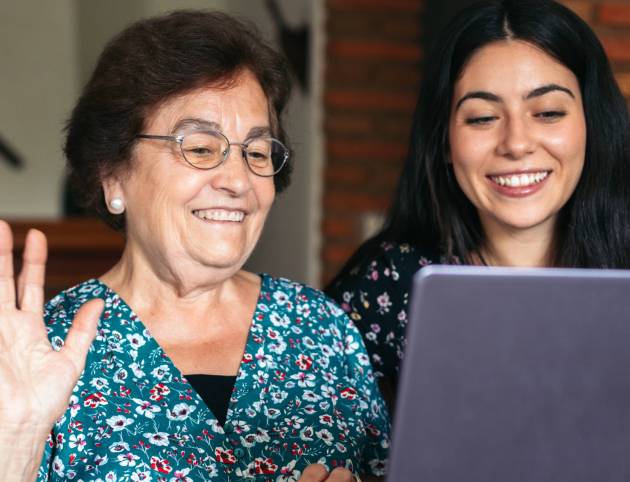 Grandmother and granddaughter making video call with laptop at home Smiling elderly woman using laptop at home making a video call together with her granddaughter. The two smile and are happy to enjoy the time together. Grandma waves to the camera